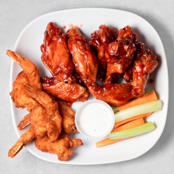 A white plate with BBQ wings, fried shrimp, ranch dressing, and carrot and celery sticks.